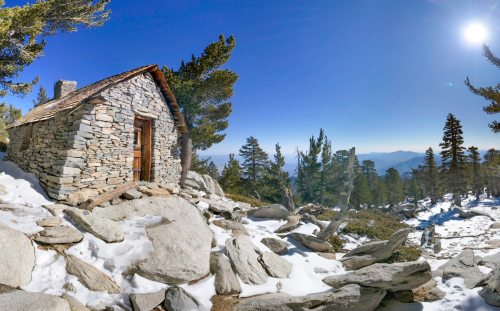 A stone cabin on a snowy landscape, surrounded by trees and mountains under a clear blue sky.