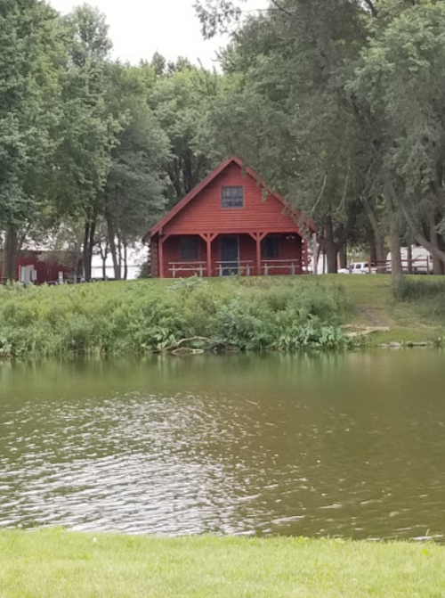 A red cabin by a calm lake, surrounded by green trees and grass.
