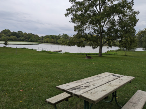A picnic table in a grassy area near a calm lake, surrounded by trees under a cloudy sky.