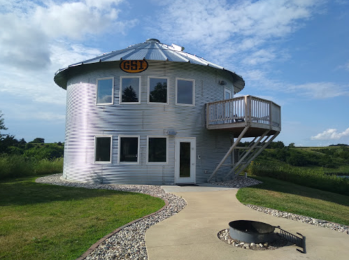 A round, silver metal house with a deck, surrounded by grass and a fire pit, under a blue sky with clouds.