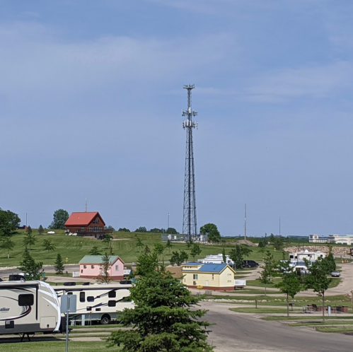 A rural landscape featuring a cell tower, a few colorful buildings, and parked RVs under a clear blue sky.