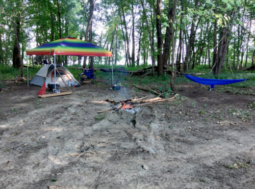 A campsite in a wooded area featuring a tent, colorful canopy, hammock, and a small fire pit on a dirt ground.
