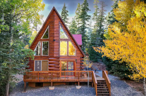 A cozy log cabin surrounded by tall trees and vibrant autumn foliage, featuring large windows and a wooden deck.