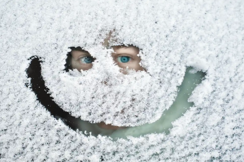 A person peeks through a snow-covered window, with only their eyes visible amidst a smiley face shape in the snow.