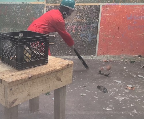A person in protective gear swings a bat to break glass bottles in a colorful, graffiti-covered room.