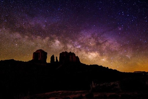 A stunning night sky filled with stars and the Milky Way, silhouetting rocky formations against a colorful horizon.