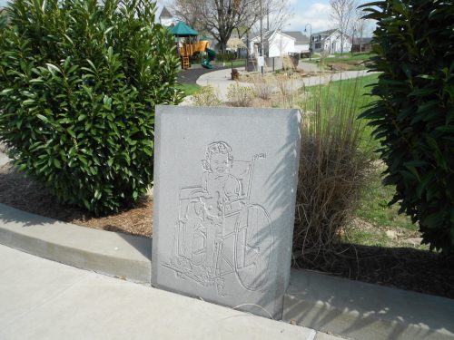 A stone sculpture of a child in a wheelchair, surrounded by greenery in a park setting.