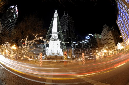 A nighttime view of a monument surrounded by city buildings, with light trails from passing vehicles.