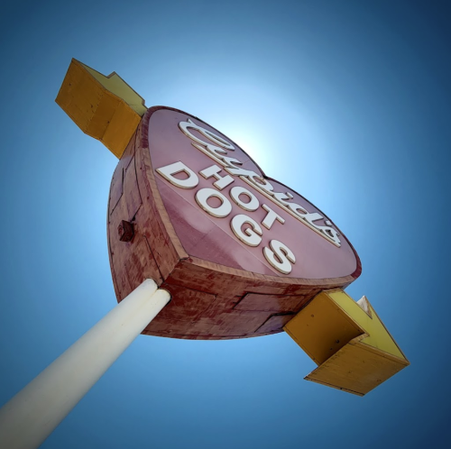 A vintage-style sign for "Crystal's Hot Dogs" with a heart shape and arrows, set against a bright blue sky.