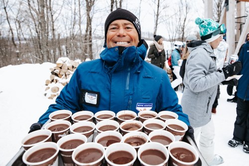 A smiling man in a blue jacket holds a tray of hot chocolate cups in a snowy outdoor setting.