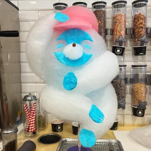 A whimsical blue and pink character made of cotton candy, playfully hugging itself against a white wall.