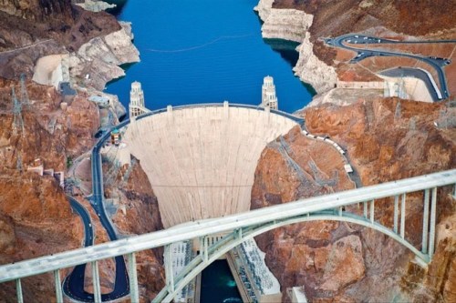 Aerial view of a large dam surrounded by rocky terrain and a blue lake, with a bridge in the foreground.
