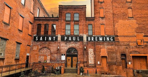 Exterior of Saint Paul Brewing, featuring red brick walls and large windows, with a sunset in the background.
