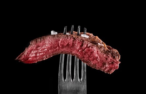 A perfectly cooked slice of steak resting on a fork, with a black background and a sprinkle of salt on top.