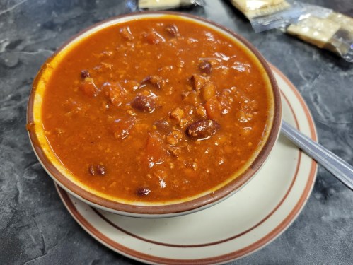 A bowl of hearty chili with beans and meat, served on a plate with a spoon beside it.