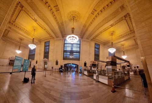 A spacious, elegantly designed train station interior with high ceilings, chandeliers, and a few people.