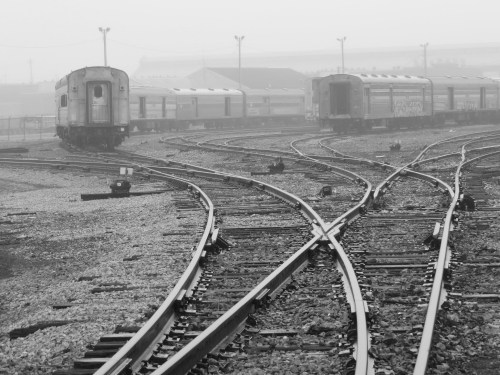 Black and white image of train tracks diverging in a foggy landscape, with abandoned train cars in the background.