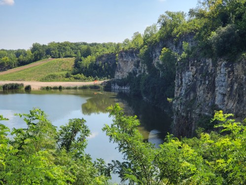 A serene landscape featuring a calm lake surrounded by lush greenery and steep rocky cliffs under a clear blue sky.