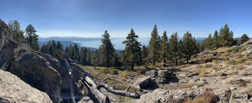 Panoramic view of a mountainous landscape with trees, rocks, and a distant lake under a clear blue sky.