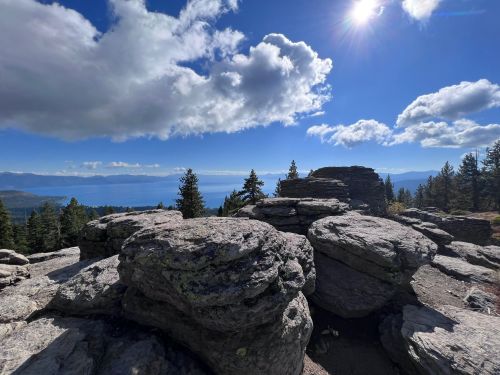 Rocky outcrop with trees and a lake in the background, under a bright sky with clouds and sunlight.
