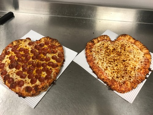 Two heart-shaped pizzas on a silver countertop: one topped with pepperoni and the other with just cheese.