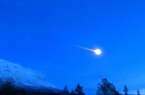 A bright meteor streaks across a clear blue sky above a snow-capped mountain landscape.
