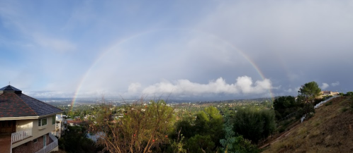 A panoramic view of a landscape with a rainbow arching over a city, surrounded by clouds and greenery.