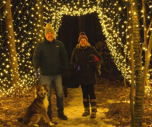A man and woman stand under a canopy of twinkling lights, holding dogs in a wooded area at night.