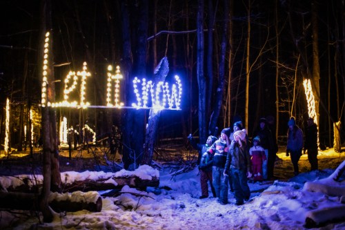 A group of people stands in a snowy forest at night, admiring a "Let it snow" light display among the trees.