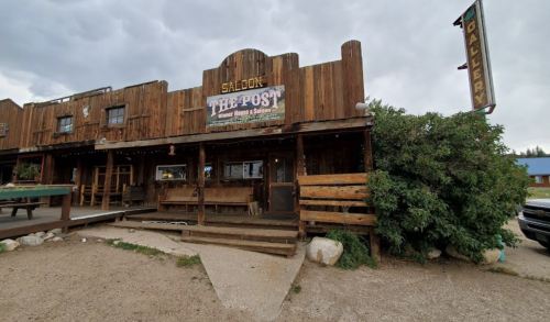 A rustic wooden saloon named "The Post" with a sign, surrounded by gravel and greenery under a cloudy sky.