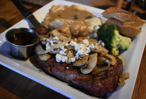 A plate featuring a grilled steak topped with onions and mushrooms, served with mashed potatoes, broccoli, and bread.