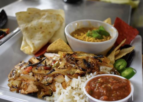 A plate of grilled chicken with rice, sautéed onions, tortillas, salsa, and a bowl of beans topped with cheese.