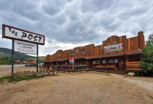 A rustic wooden building with a sign reading "The Post" and a saloon, set against a cloudy sky.