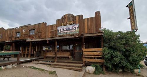 A rustic wooden saloon named "The Post" with a sign and outdoor seating, set against a cloudy sky.