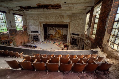 Abandoned theater interior with worn seats, peeling walls, and a cluttered stage, illuminated by natural light through windows.