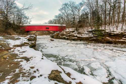 A red covered bridge spans a frozen river, surrounded by snow-covered rocks and bare trees in a winter landscape.