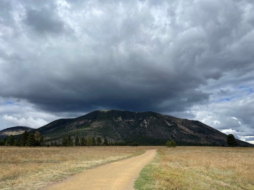 A winding dirt path leads through a grassy field towards a mountain under a dramatic, cloudy sky.