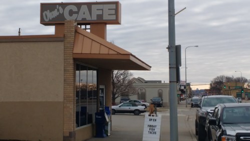 Exterior of Olie's Cafe with a sign advertising Friday fish tacos, parked cars, and a cloudy sky in the background.