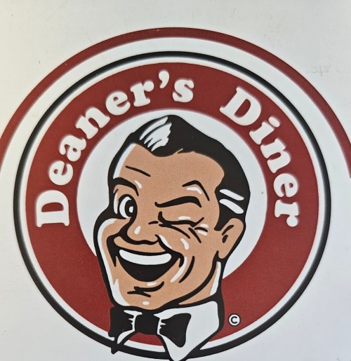 Logo of Deaner's Diner featuring a smiling, winking character in a red and white circular design.