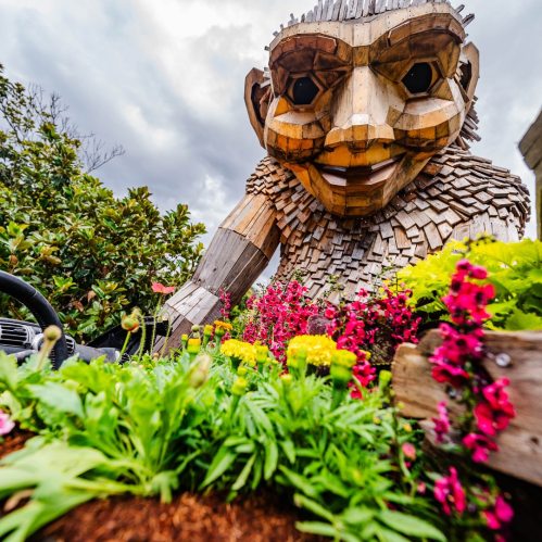 A large wooden sculpture of a smiling child surrounded by colorful flowers and greenery under a cloudy sky.