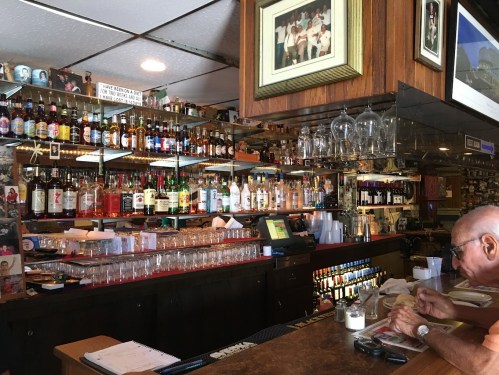 A cozy bar with a wooden counter, shelves lined with various bottles, and a man sitting at the bar.
