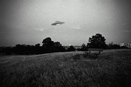 A black and white image of a UFO hovering above a city skyline, with a bench in a grassy field in the foreground.