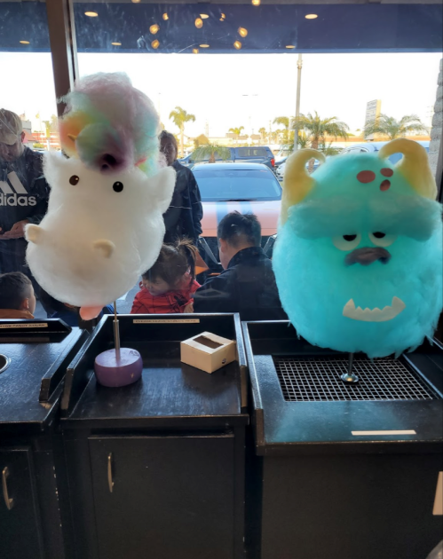 Two colorful cotton candy creations resembling a fluffy white creature and a blue monster, displayed in a shop window.