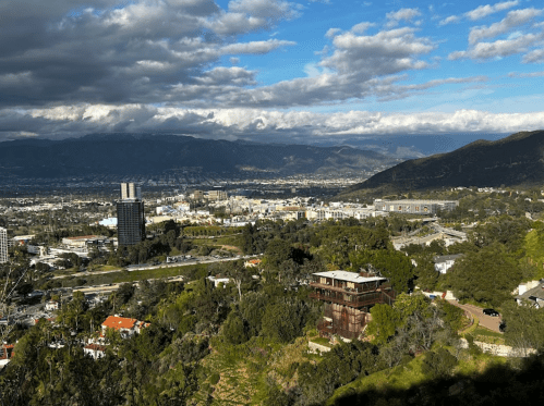 A panoramic view of a cityscape with mountains in the background, featuring buildings and greenery in the foreground.