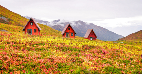 Three red cabins sit amidst vibrant autumn foliage on a mountainous landscape under a cloudy sky.