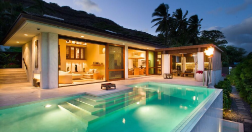 A modern villa with a pool, surrounded by palm trees, illuminated at dusk. Cozy indoor and outdoor living spaces visible.