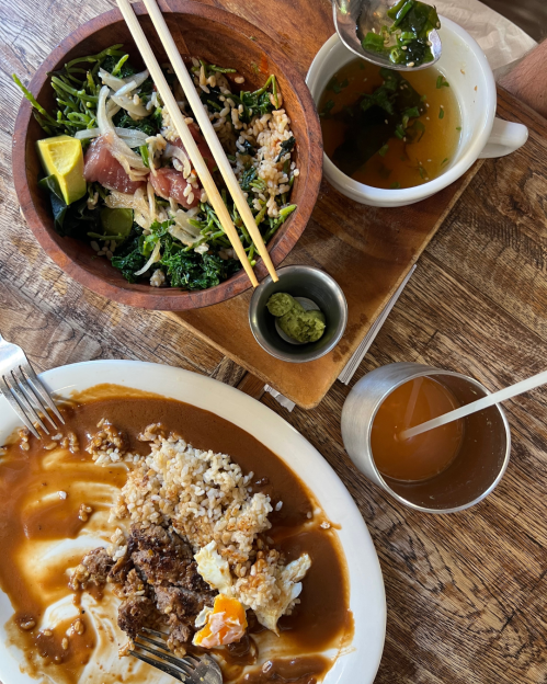 A wooden table with a plate of rice and meat, a salad bowl, soup, and a drink, all arranged for a meal.