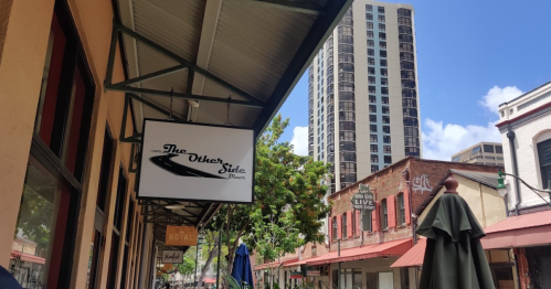 A street view featuring a sign for "The Other Side" restaurant, with tall buildings and blue sky in the background.