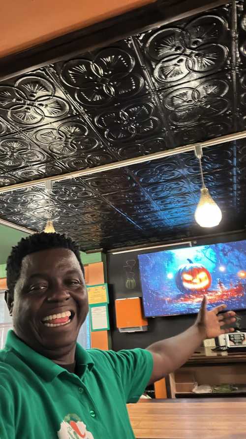 A smiling man in a green shirt gestures towards a TV displaying a Halloween-themed image of a pumpkin.