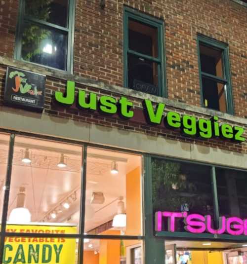 Signage for "Just Veggiez" restaurant and "IT'SUGAR" candy store, illuminated at night.